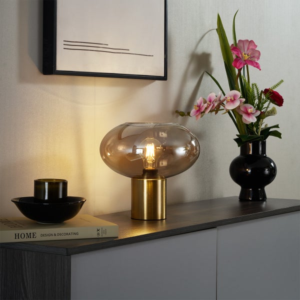Moderne Tischleuchte mit goldfarbenem Fuß und rauchgrauem Glasschirm auf einem Sideboard neben einer Blumenvase und Dekoration.