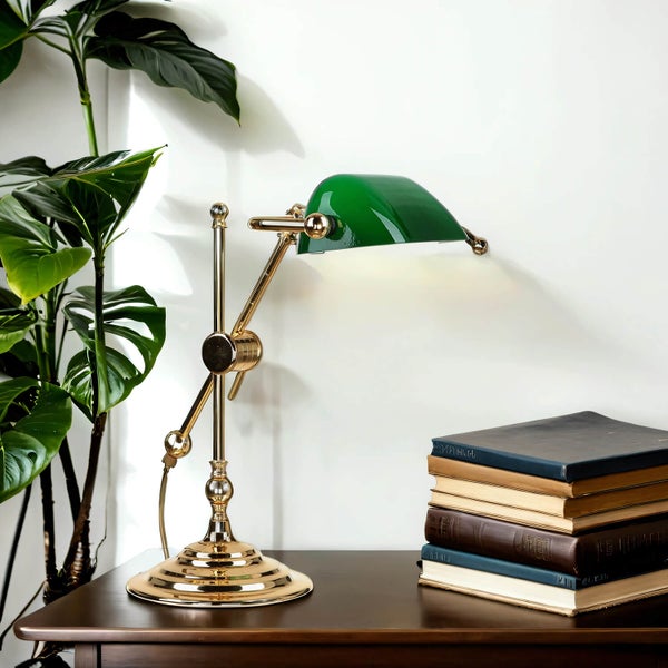 Tischleuchte mit grüner Abdeckung und goldenem Gestell auf einem Tisch mit Büchern und einer Pflanze.