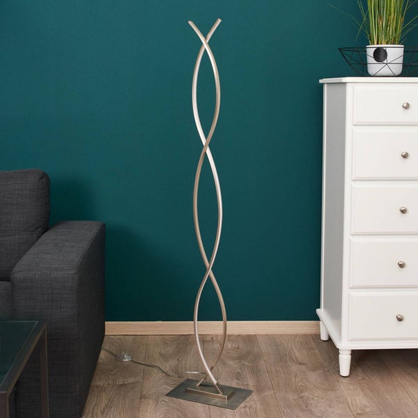 Moderne LED-Stehleuchte aus Metall mit zwei geschwungenen Elementen in einem Wohnzimmer neben einem grauen Sofa und einer weißen Kommode.