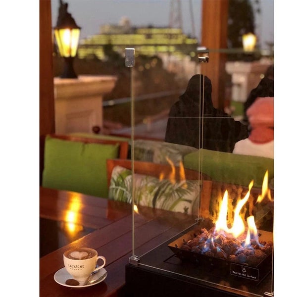 Moderner Tischkamin mit Glasscheiben und brennenden Flammen auf einem Holztisch neben einer Tasse Kaffee in einer gemütlichen Außenlounge.