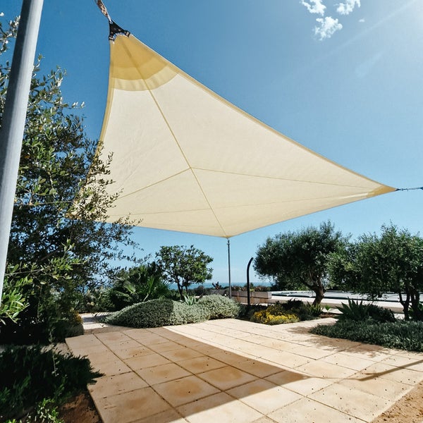 Dreieckiges Sonnensegel gespannt über einer hellen Terrasse im Garten, umgeben von Pflanzen und Bäumen an einem sonnigen Tag.