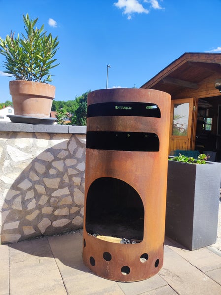 Zylindrischer Gartenkamin aus Cortenstahl auf einer Terrasse neben einer Steinmauer und Pflanzgefäßen.