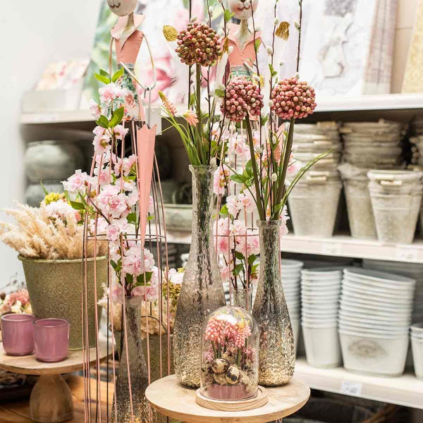 Frühlingsdekoration mit künstlichen rosa Blüten in strukturierten Glasvasen, einer Glasglocke mit Eiern und verschiedenen Übertöpfen im Regal.