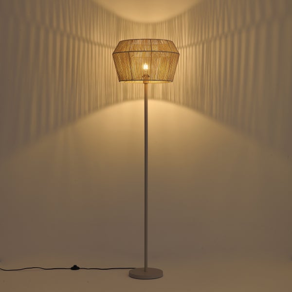 Eingeschaltete Stehleuchte mit gewebtem Lampenschirm aus Naturfaser, die ein warmes Licht und dekorative Streifenschatten an die Wand wirft.