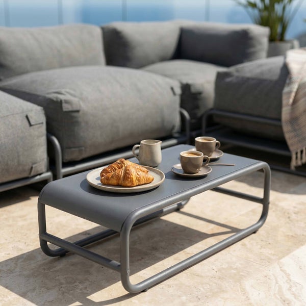 Moderner Garten-Loungetisch aus Metall in Anthrazit auf einer hellen Terrasse vor einem grauen Lounge-Sofa mit Kaffee und Croissants.