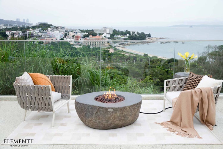 Moderne Terrasse mit Elementi Gas-Feuertisch in Steinoptik, Loungesesseln und Blick auf das Meer und eine Küstenstadt.