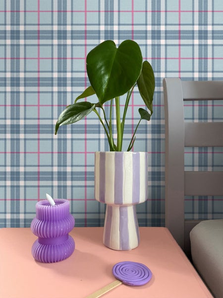 Innenraumszene mit karierter Tapete einer Monstera-Pflanze in einem gestreiften Keramiktopf und einer lila Kerze auf einem rosa Tisch.