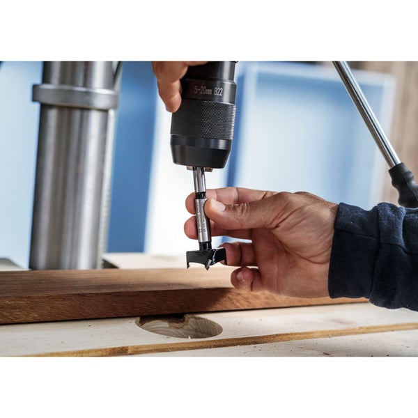 Eine Hand bedient eine Tischbohrmaschine mit einem Forstnerbohrer, um ein Loch in ein Holzbrett zu bohren.