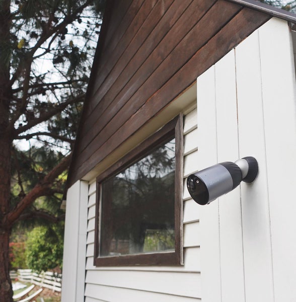 EZVIZ Überwachungskamera montiert an einer weißen Holzwand eines Gartenhauses neben einem Fenster.