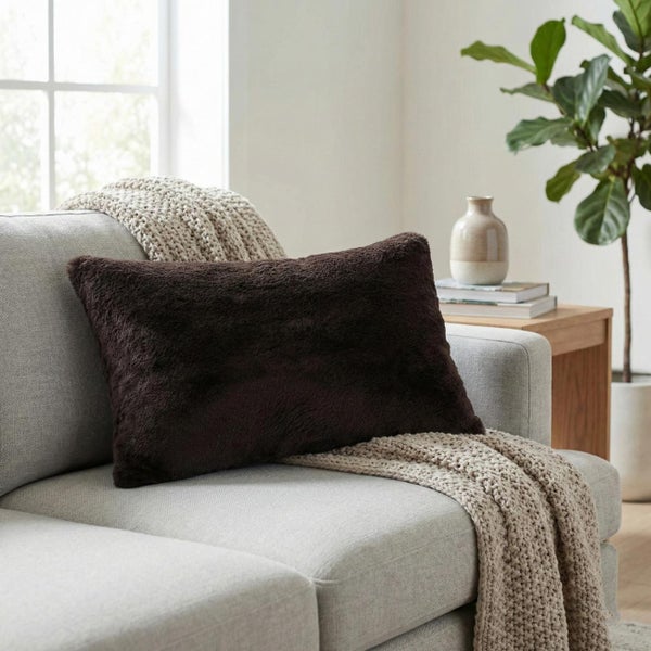 Rechteckiges Dekokissen in Dunkelbraun mit Kunstfell-Optik auf einem hellgrauen Sofa in einem hellen Wohnzimmer.