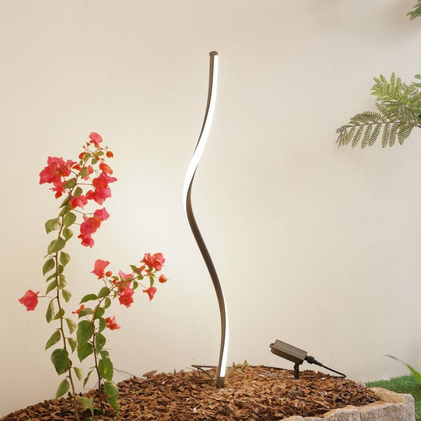 Geschwungene LED-Gartenleuchte in einem mit Rindenmulch bedeckten Beet neben einer blühenden Pflanze.