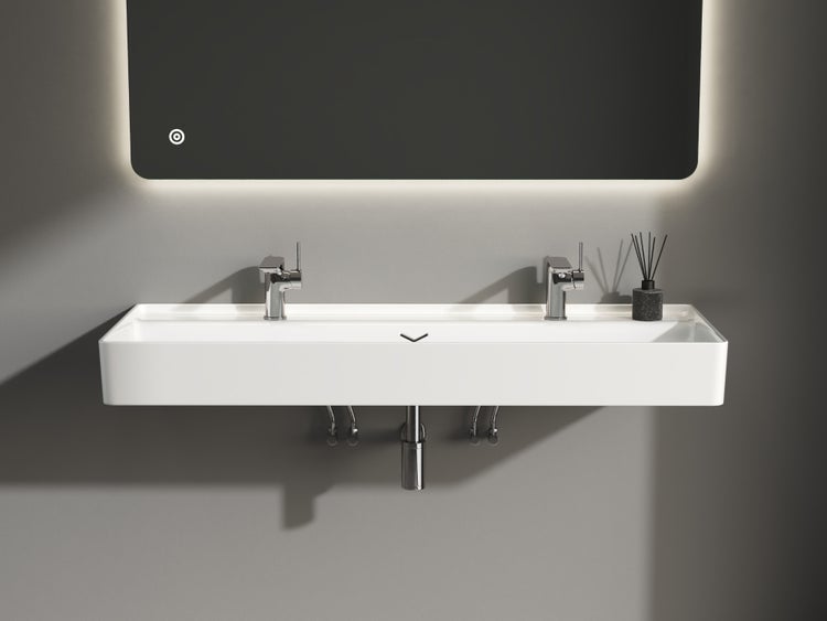 Modernes Badezimmer mit breitem weißem Wandwaschbecken, zwei verchromten Armaturen und einem hinterleuchteten Spiegel darüber.