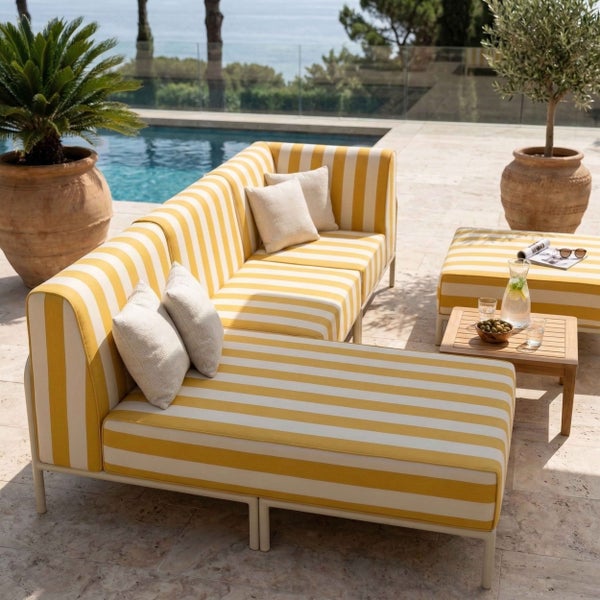Moderne Gartenlounge mit gelb-weiß gestreiftem Ecksofa, Hocker, Holzbeistelltisch und Pflanzen an einem Pool auf einer hellen Steinterrasse.