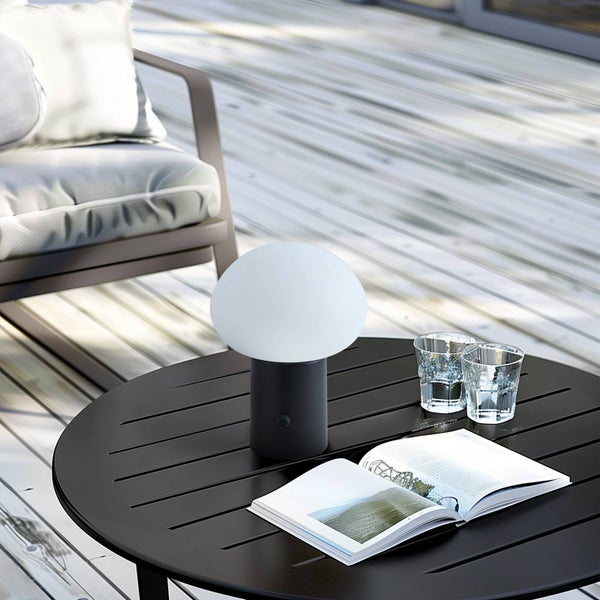 Moderne tragbare Tischleuchte mit weißem Schirm auf schwarzem Metalltisch auf einer hellen Holzterrasse neben einem grauen Gartensessel.