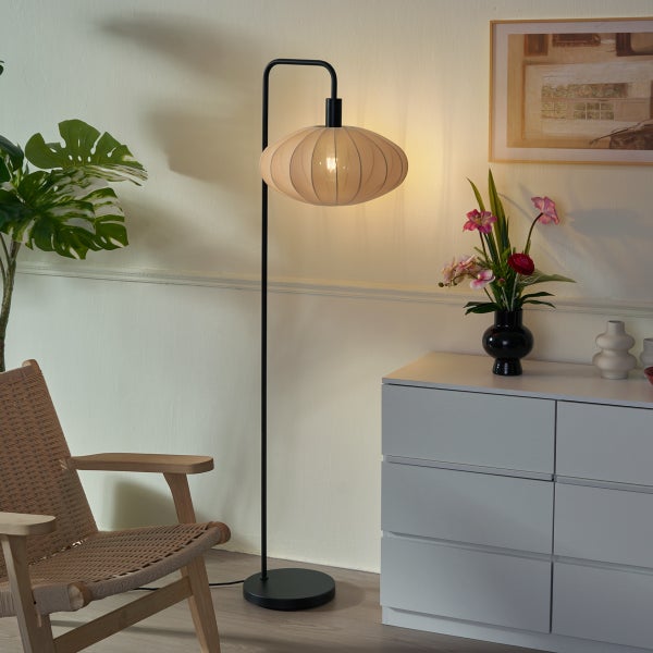 Moderne Stehleuchte mit schwarzem Metallgestell und beigem Stoffschirm in einem hellen Wohnzimmer neben einem Holzstuhl und einer weißen Kommode.