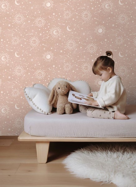 Helles Kinderzimmer mit Tapete mit Sonnen, Monden und Sternen, ein Kind liest ein Buch auf einem Holzbett neben Kissen und einem Stoffhasen.