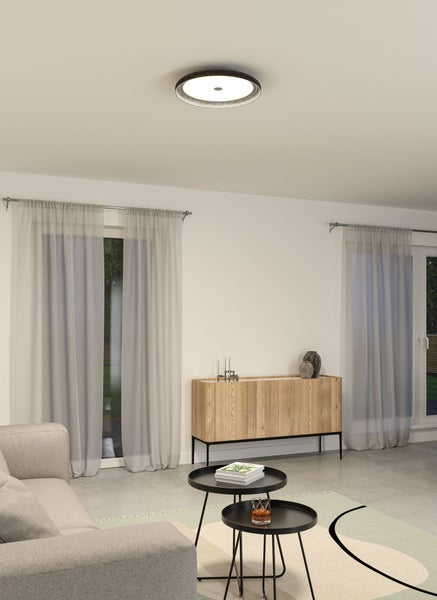 Modernes, helles Wohnzimmer mit runder Deckenleuchte, einem Sideboard aus Holz, zwei schwarzen Couchtischen und hellen Vorhängen.