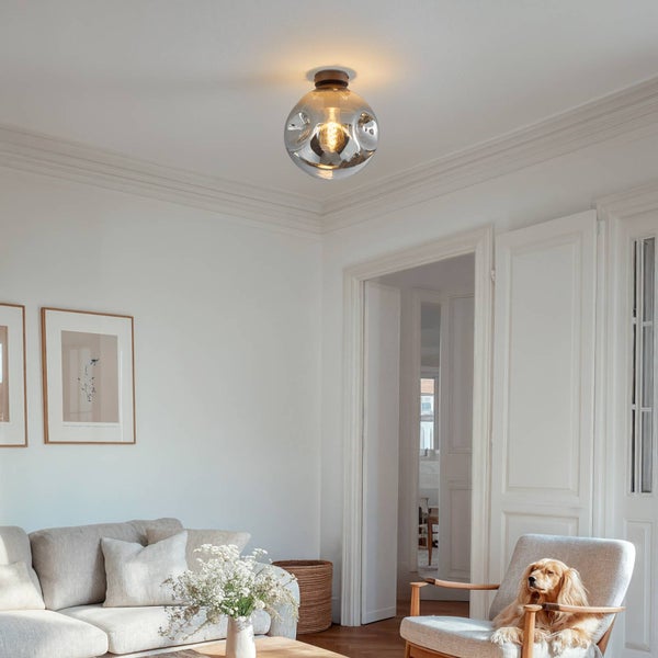 Helles, geräumiges Wohnzimmer mit einer modernen Deckenleuchte aus Rauchglas. Die Szene umfasst ein graues Sofa, einen Sessel und weiße Wände.