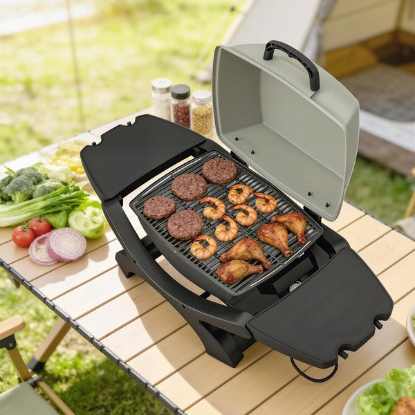 Tragbarer Tischgrill auf einem Holztisch im Freien mit Fleisch, Garnelen und Gemüse in einer hellen Camping-Umgebung.