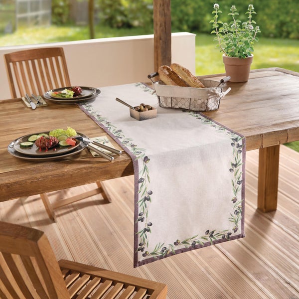 Tischläufer mit Olivenzweig Muster auf einem rustikalen Holztisch im Garten, dekoriert mit Tellern, Brot und Besteck.