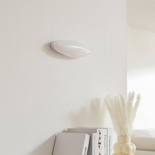 Ovale Wandleuchte in Weiß in einem modern gestalteten Wohnraum über einer Dekoration aus Büchern und einer Vase mit Trockenblumen.
