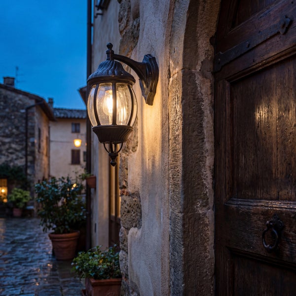 Klassische Außenwandleuchte in Laternenform an einer hellen Hauswand neben einer dunklen Holztür in einer abendlichen Gasse.