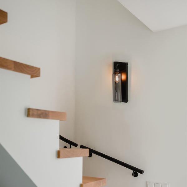 Moderne Wandleuchte mit zylindrischem Glasschirm an weißer Wand im hellen Treppenhaus mit Holzstufen und schwarzem Handlauf.