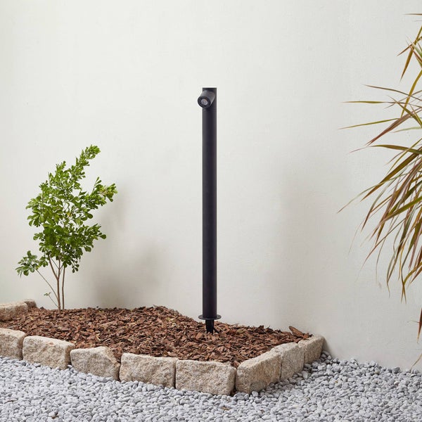 Schwarze Pollerleuchte für den Garten in einem Beet mit Rindenmulch und Steineinfassung.