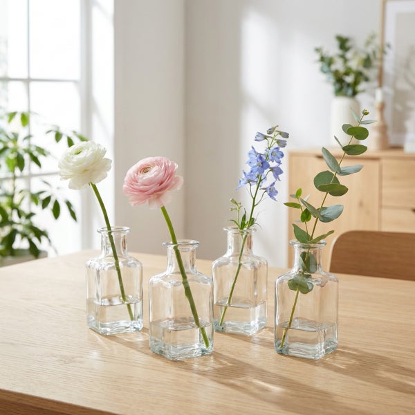 Vier kleine, klare Glasvasen in quadratischer Form mit Blumen und Eukalyptuszweigen dekoriert auf einem Holztisch.