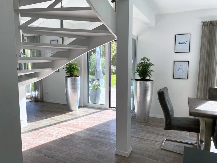 Moderner heller Innenraum mit weißer Wendeltreppe, Boden in Holzoptik und großen silbernen Pflanzkübeln neben einer Glastür zum Garten.