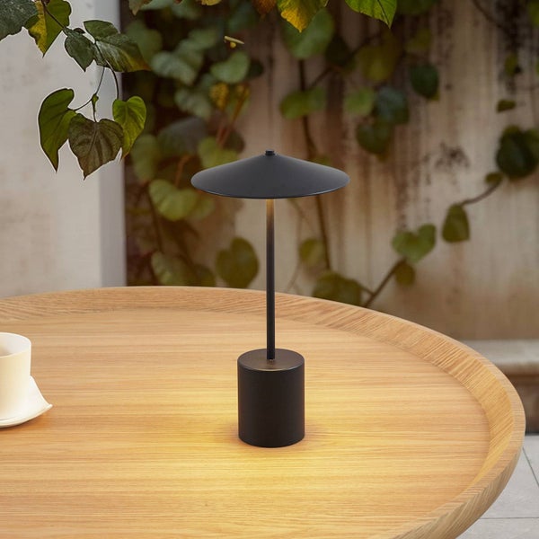 Tragbare LED-Tischleuchte in Mattschwarz auf einem runden Holztisch im Außenbereich mit atmosphärischer Beleuchtung.