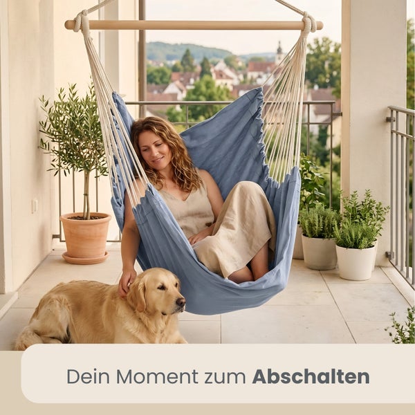 Eine Frau entspannt in einem blauen Hängesessel auf einem hellen Balkon mit Pflanzen und einem Hund.