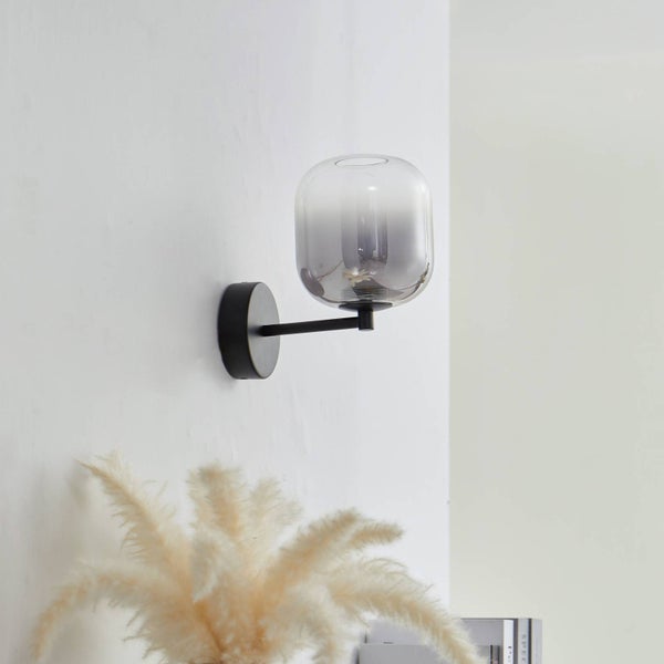 Moderne Wandleuchte mit rauchgrauem Glasschirm und schwarzer Metallhalterung an einer hellen Wand mit dekorativem Pampasgras im Vordergrund.