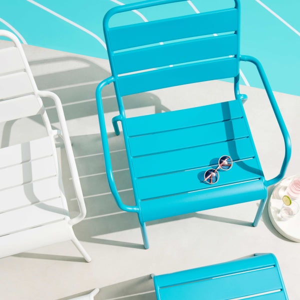 Gartenstühle aus Metall mit Lattenstruktur an einem Pool, ein türkisfarbener Sessel mit Sonnenbrille und ein weißer Stuhl im hellen Sonnenlicht.