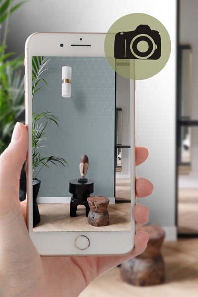 Eine Person hält ein Mobiltelefon, das ein Zimmer mit einer geometrischen Tapete, einer Wandleuchte und dekorativen Hockern zeigt.