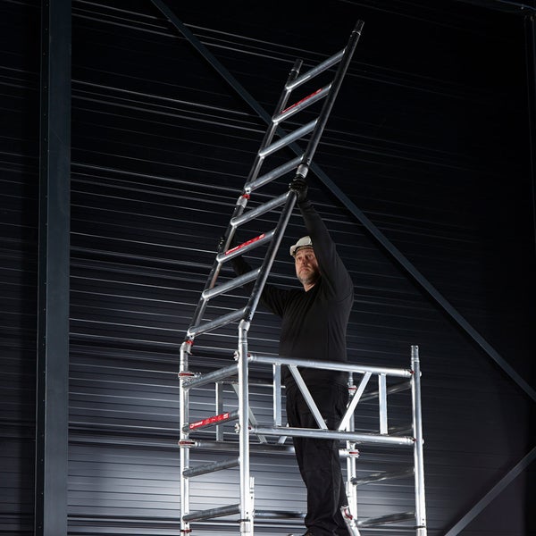 Mann montiert ein Gerüst mit Leiteraufstieg in einer Halle.