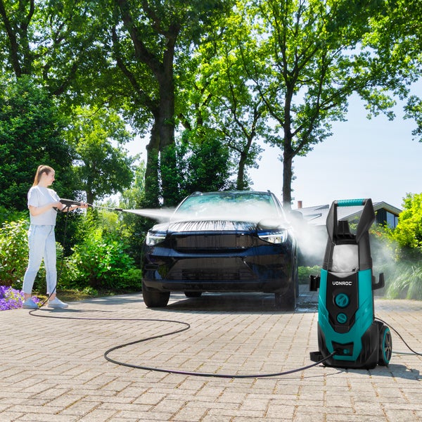 Eine Frau reinigt ein schwarzes Auto mit einem Vonroc Hochdruckreiniger auf einer gepflasterten Auffahrt im sonnigen Außenbereich.