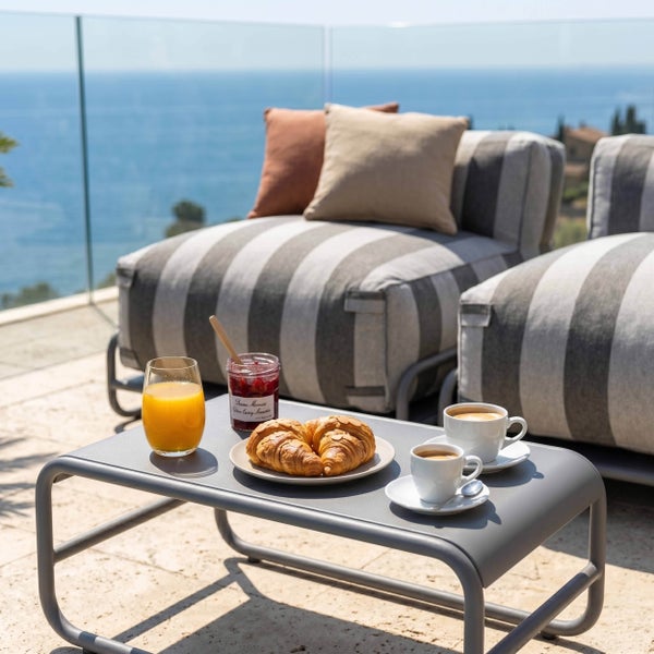 Heller Loungebereich auf einer Terrasse mit Meerblick, gestreiften Sesseln, Kissen und einem Beistelltisch aus Metall mit Frühstück.