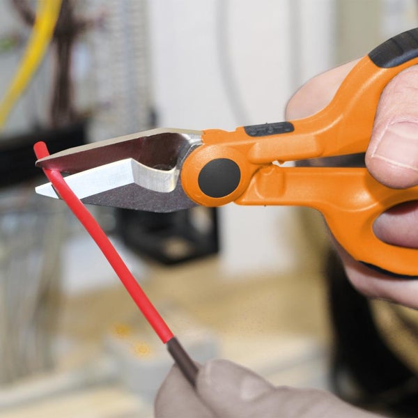 Elektrikerschere mit orangefarbenem Griff schneidet ein rotes Kabel in einer hellen Elektroinstallation.
