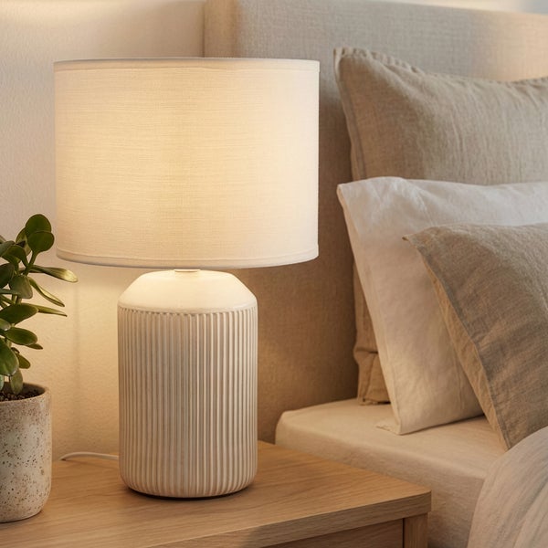 Tischleuchte mit geriffeltem Keramikfuß und Stoffschirm auf einem Holznachttisch neben einem Bett mit Kissen in einem hellen Schlafzimmer.