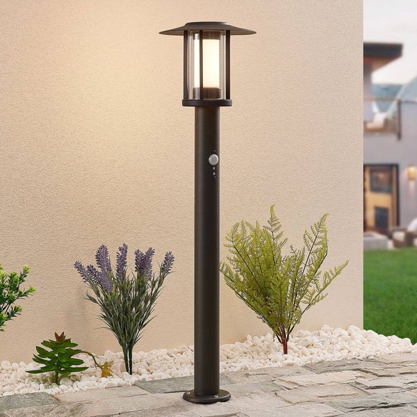 Moderne Wegeleuchte aus Metall mit Bewegungsmelder, beleuchtet in einem Gartenbeet mit Kies, Lavendel und Farn vor einer hellen Wand.