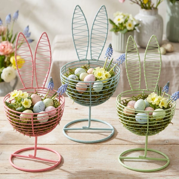 Drei dekorative Osterhasen-Schalen aus Metall mit Hasenohren zur Osterdekoration gefüllt mit Moos, Ostereiern und Frühlingsblumen auf einem Tisch.