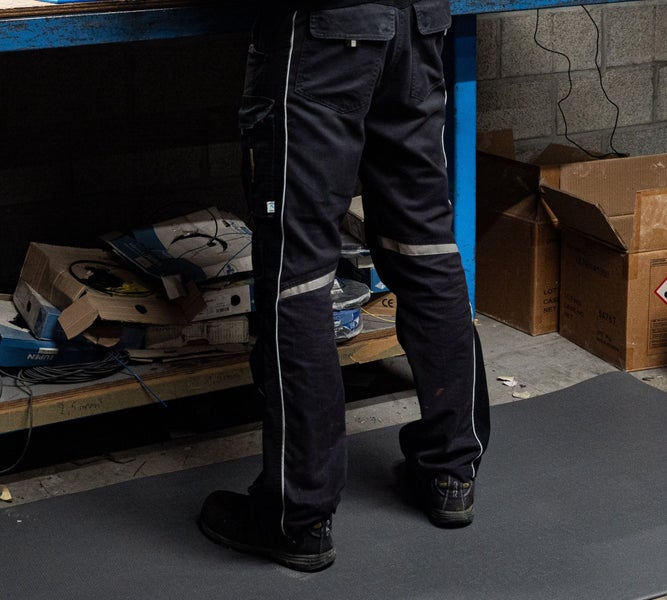 Person in einer Arbeitshose mit Reflexstreifen und funktionalen Taschen in einer Werkstattumgebung.
