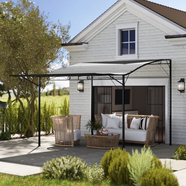 Wand-Pergola mit schwarzem Metallgestell und hellem Stoffdach über einer Terrasse mit Korbmöbeln vor einem weißen Haus.