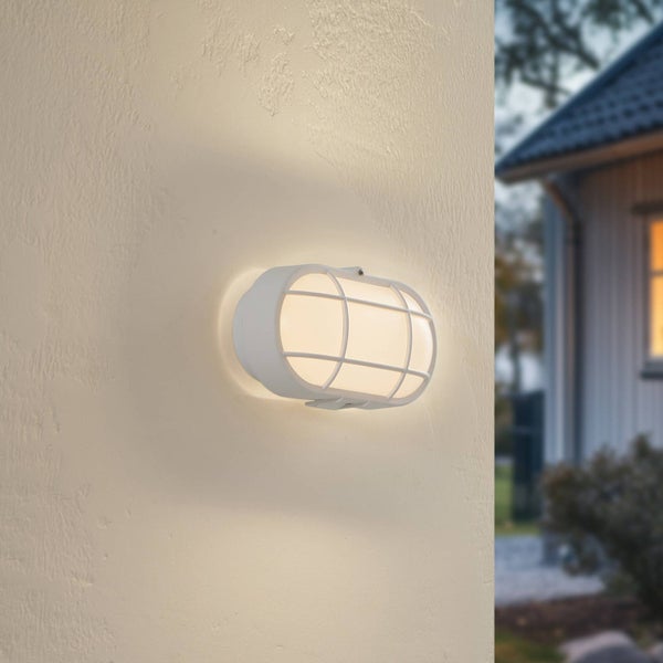 Beleuchtete ovale Außenwandleuchte mit weißem Schutzkorb an einer strukturierten Fassade.
