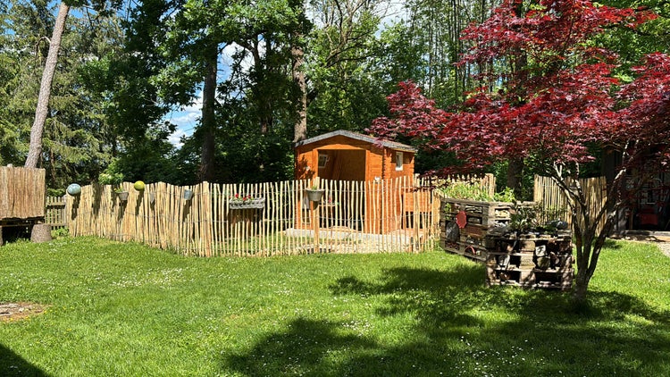 Gartenhaus aus Holz hinter einem Staketenzaun in einem hellen Garten mit Insektenhotels aus Paletten und einem roten Ahorn.