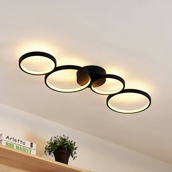 Moderne LED-Deckenleuchte mit vier schwarzen, ineinandergreifenden Ringen, warmweißes Licht, montiert an der Decke über einem Regal.