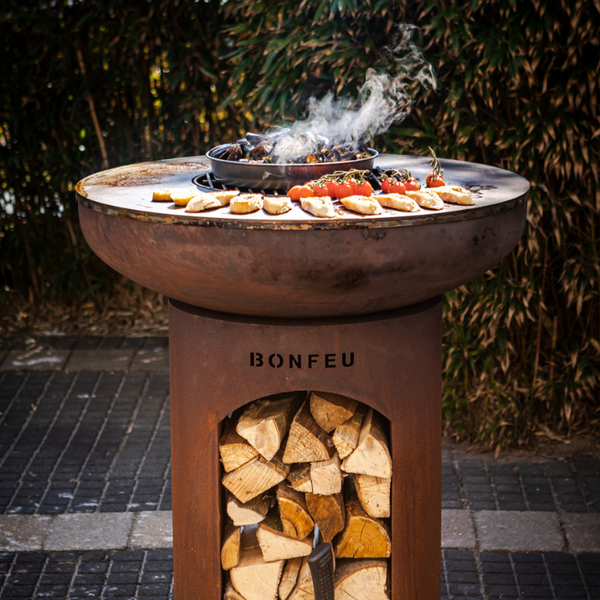 BonFeu Außengrill aus Cortenstahl mit Holzaufbewahrung, bestückt mit Muscheln, Tomaten und Brot auf einer runden Grillplatte.