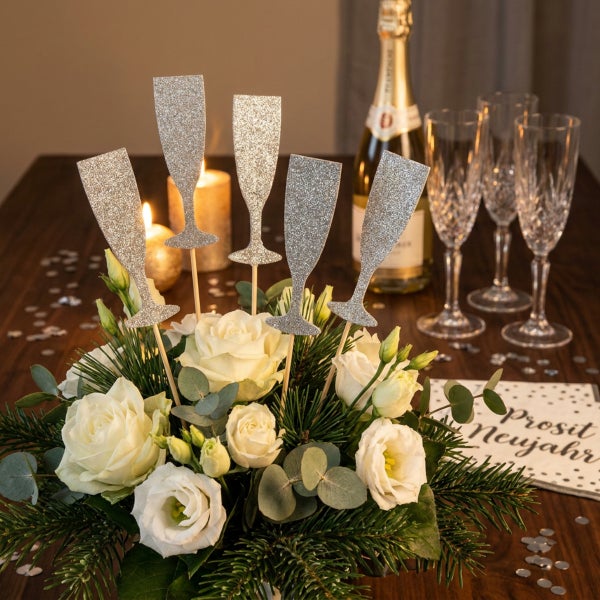 Silberne Sektglas Stecker als Tischdekoration in einem Blumenstrauß mit weißen Rosen und Tannengrün für eine Silvesterparty.