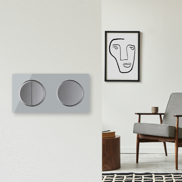 Doppellichtschalter in moderner Optik an einer Wand vor einem stilvoll eingerichteten Wohnbereich mit Sessel und abstraktem Bild.